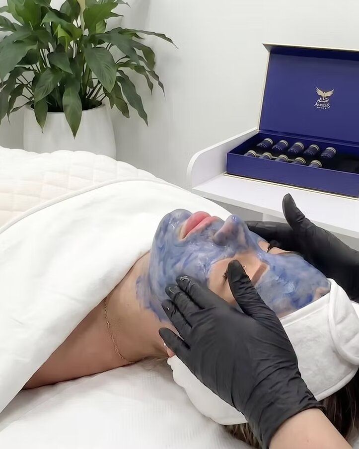 Behandlerin in schwarzen Handschuhen massiert die blaue Bio-Microneedling-Maske in das Gesicht einer Klientin ein